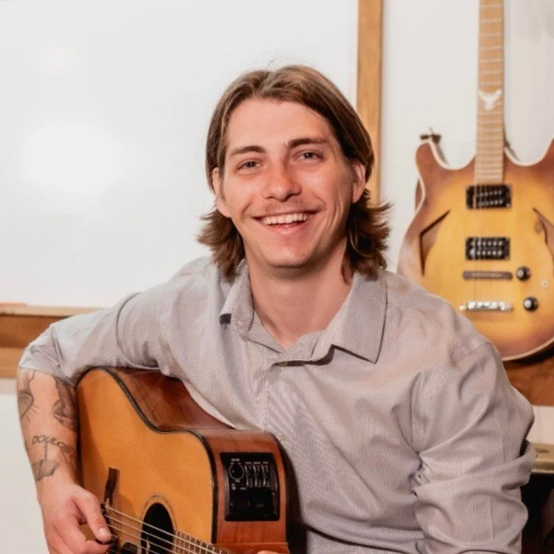 A young man with shoulder-length brown hair smiling while holding an acoustic guitar in a room with a whiteboard and an electric guitar hanging on the wall behind him.
