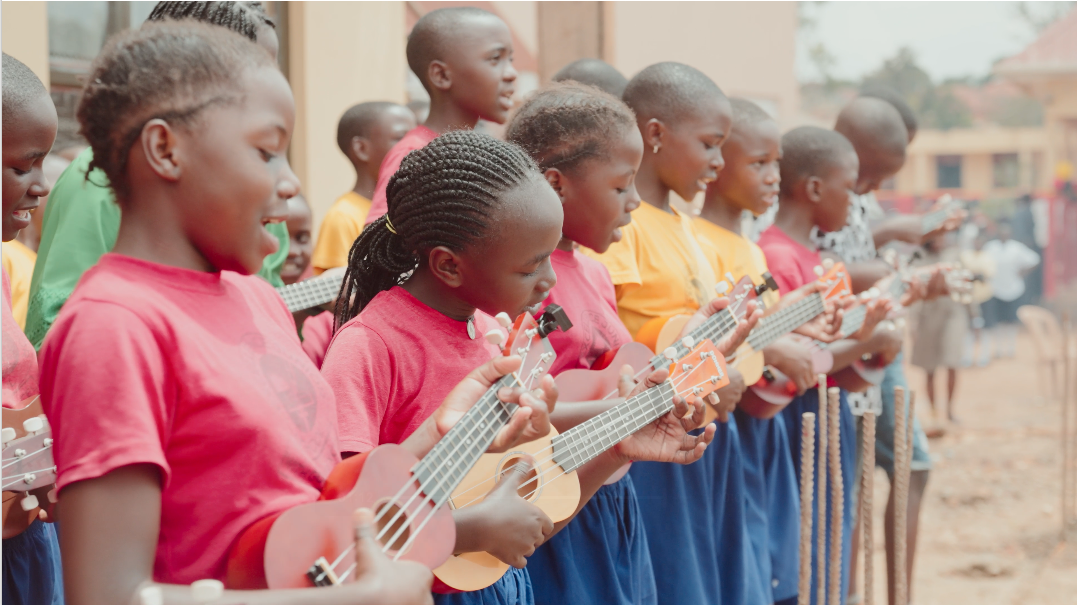 Group of young children in colorful shirts, playing small ukuleles outdoors.