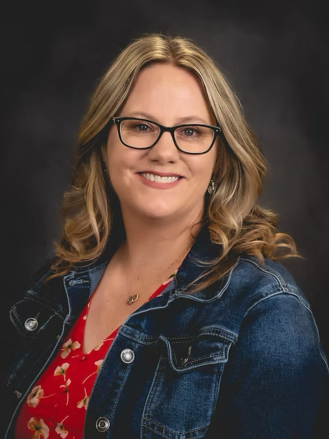 Portrait of a woman with glasses, blond hair, smiling, wearing a denim jacket over a red floral top.