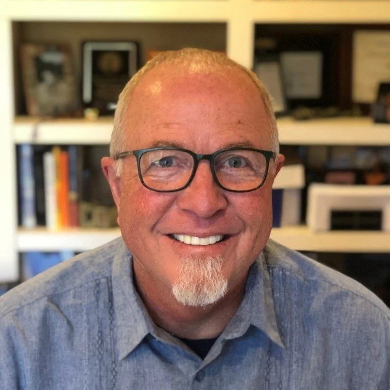A smiling man with glasses and a goatee in a gray shirt, with a background of books and shelves.