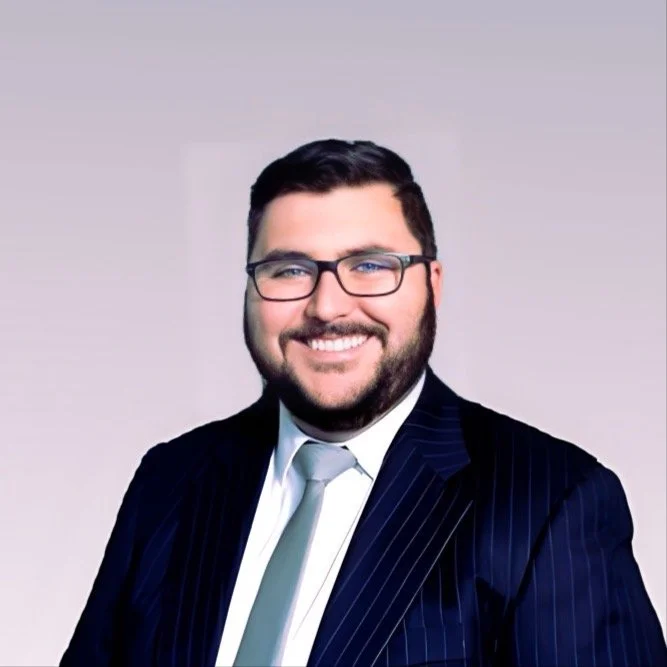 A smiling man with glasses and a beard, dressed in a dark pinstripe suit, white shirt, and light-colored tie, posed against a plain light background.