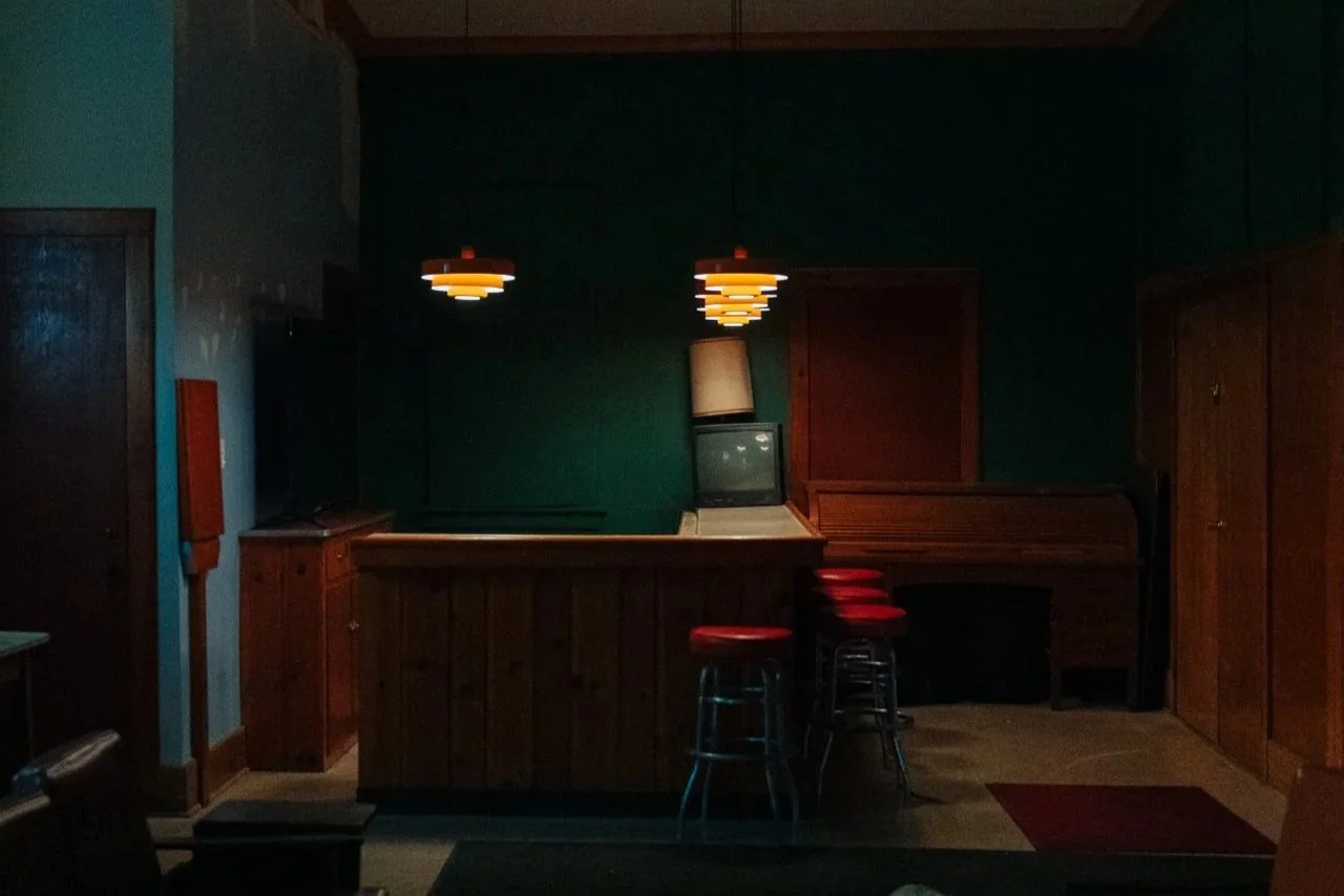 A dimly lit interior of a bar or diner with a wooden counter, three red bar stools, a small vintage television, a tall lamp, and two hanging orange and white pendant lights.