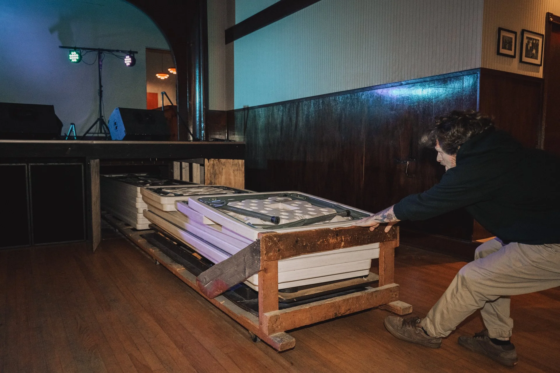 A person pushing a cart loaded with white folding tables and a metal frame in a dimly lit venue with a stage area and blue and purple lighting.