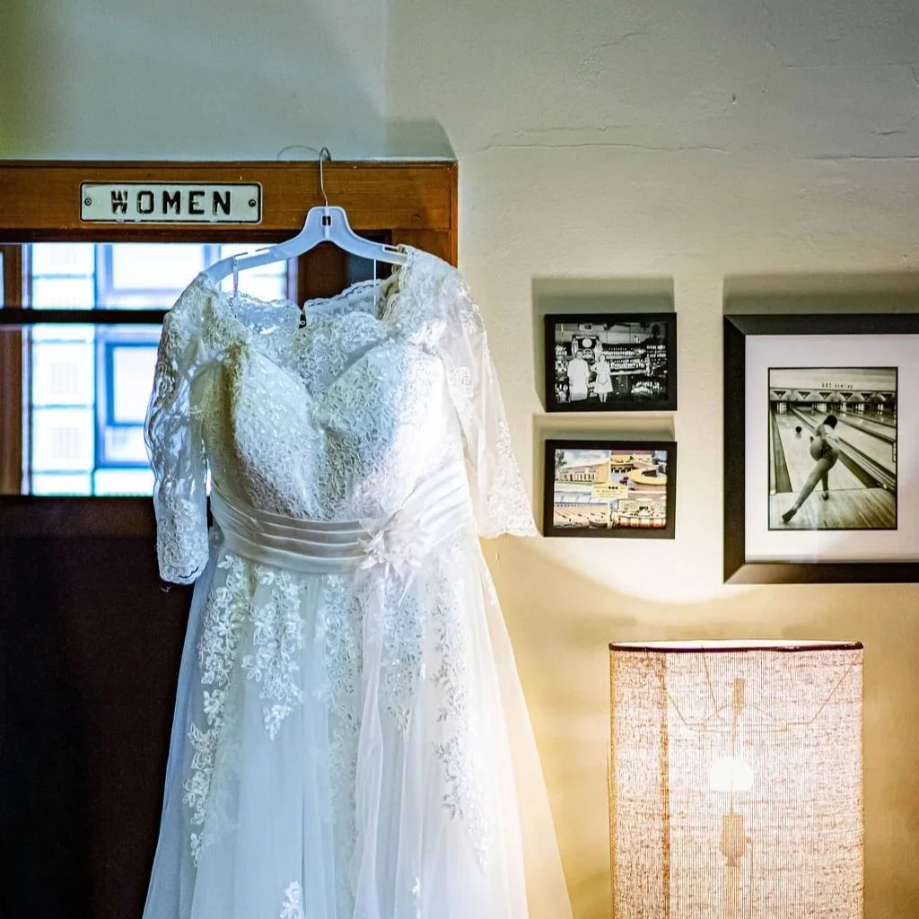 A white wedding lace dress hanging on a white hanger in front of a mirror in a room with framed photographs and a lamp.
