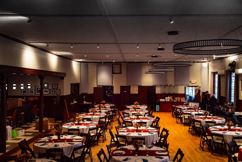 A restaurant with round tables set for dining, decorated with flowers, napkins, and utensils. There are people at the counters and the room has a cozy, dimly lit ambiance.