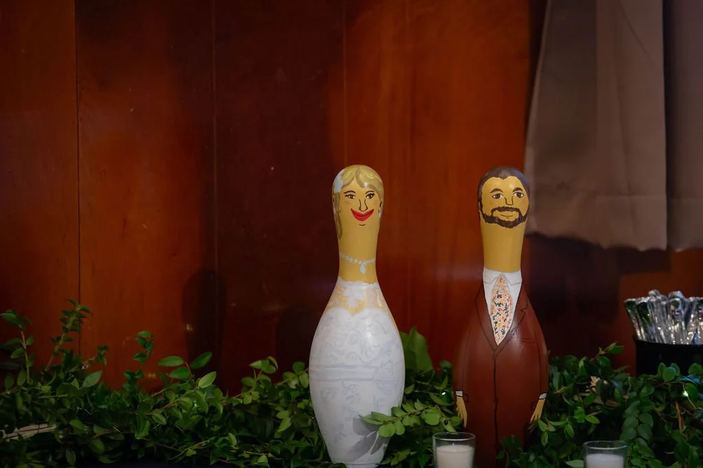 Wooden wall with two painted decorative figurines of a woman in a wedding dress and a man in a suit surrounded by greenery and candles.