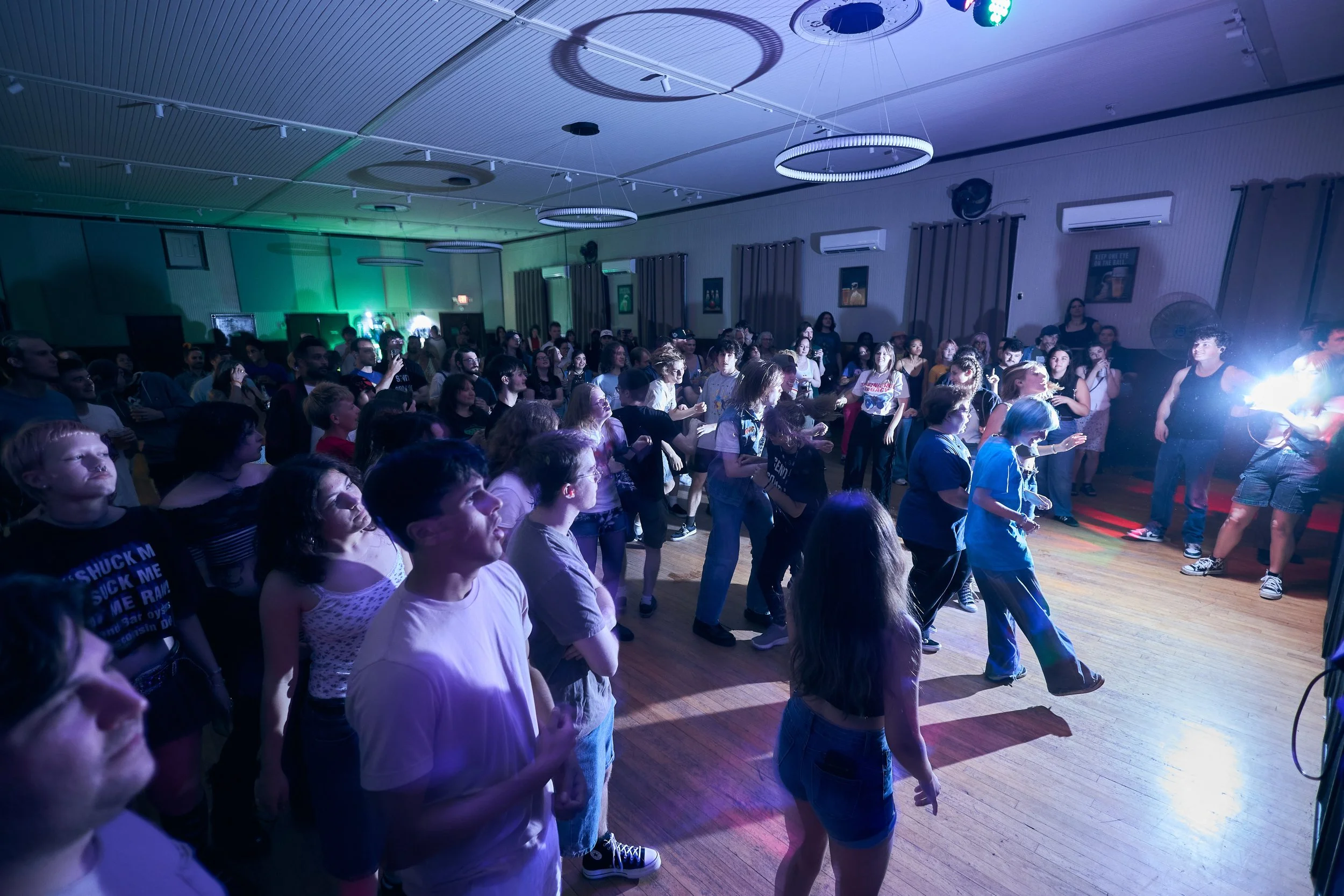 A large group of people attending a concert or dance event in a dimly lit indoor venue with colorful stage lights. Some are dancing while others watch the performance or socialize.