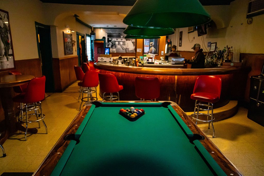 Interior of a bar with a pool table in the foreground and bar stools around the bar counter in the background.
