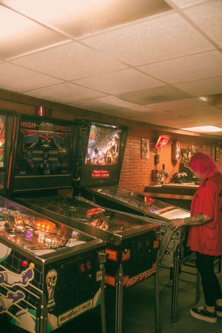 A person with pink hair playing pinball at a vintage arcade with brick walls and arcade posters.