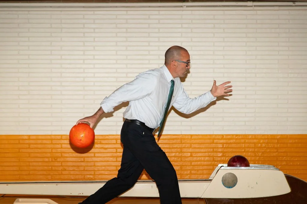 A man in business attire holding a bowling ball and preparing to bowl in a bowling alley.