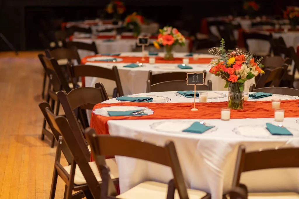 Elegant banquet setup with round tables covered in white tablecloths and orange table runners, decorated with colorful flower arrangements and teal napkins. Small candles are placed on the tables, and table number signs are displayed, suggesting a fo