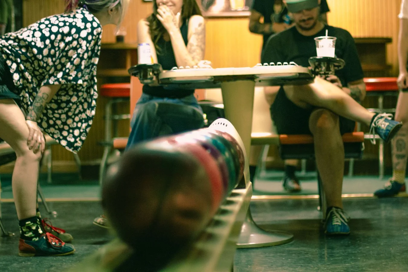 People playing a video game at a bar, with a focus on a person on the left leaning forward and another person sitting at the table with drinks and tattoos.