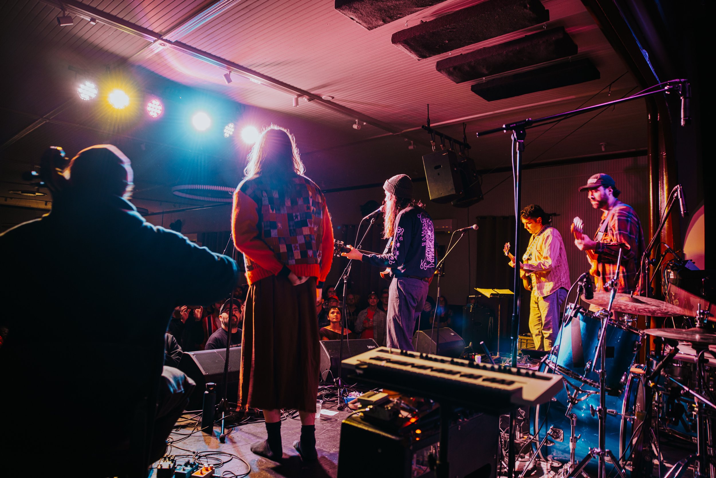Band performing on stage with colorful lights, audience watching, musical instruments including keyboard and drums visible.