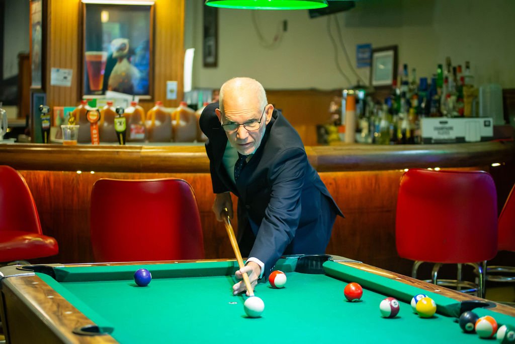 A man in a suit playing pool on a green billiard table in a bar or pub, with red chairs and a bar in the background.