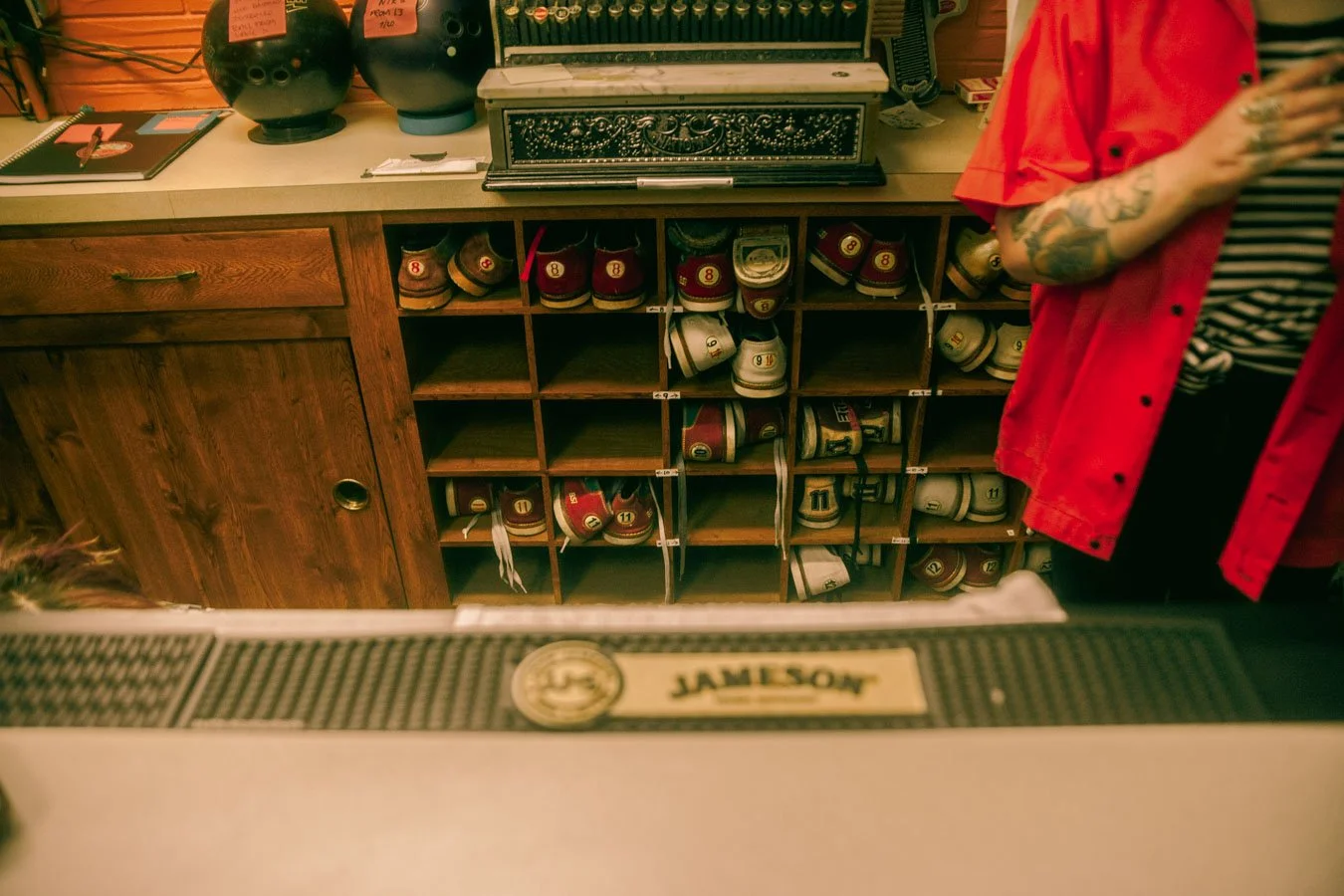 A wooden cubby shelf filled with bowling shoes, with a counter above. On the counter, there are two large bowls, a notepad, and other items. Part of a person wearing a red jacket and striped shirt is visible on the right side.