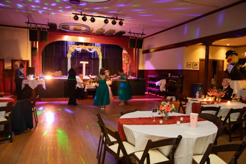 People dancing and socializing in a decorated banquet hall with a stage, tables with flowers, and a large mascot in the corner.