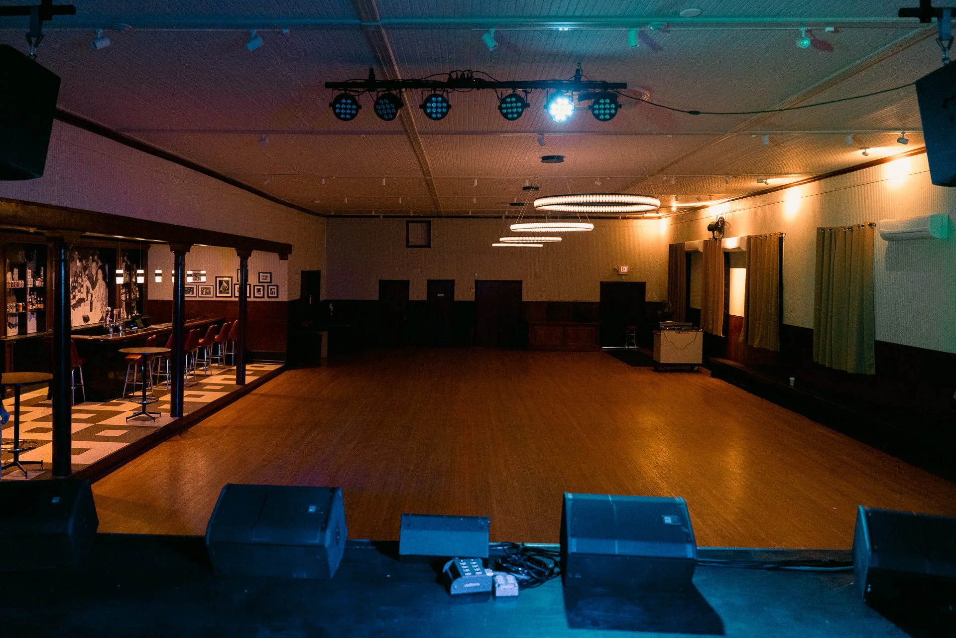 Empty stage with speakers and equipment in a dimly lit performance or dance hall, with a bar area along the left side.