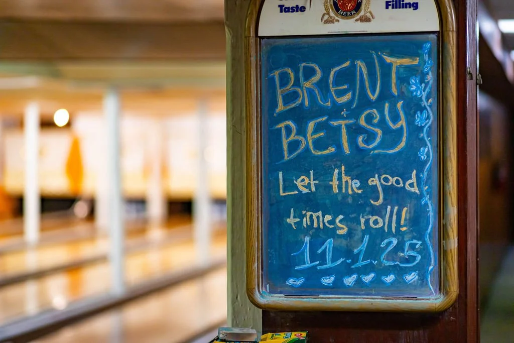 A chalkboard sign with the words 'Brent & Betsy Let the good times roll!! 11.12.25' written on it, decorated with small hearts at the bottom. The sign is attached to a wooden post at a store or restaurant.