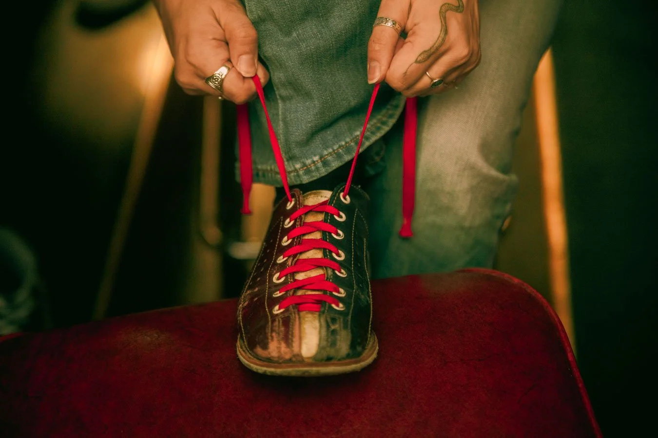 Person tying red shoelaces on a black and brown worn-out sneaker, standing on a red surface.