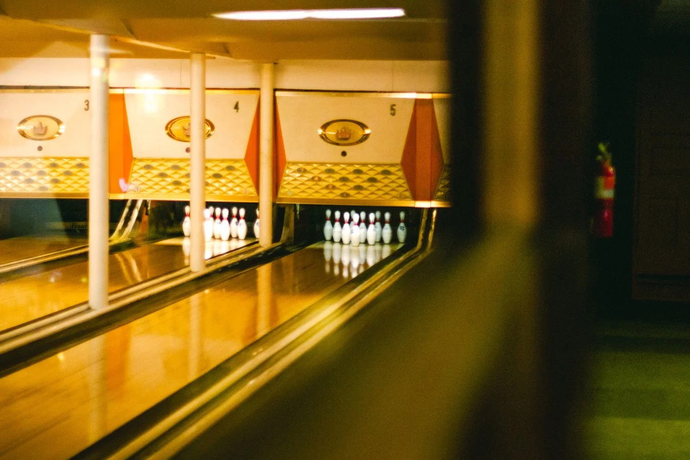 Bowling alley with pins set up at the end of the lane, lane number signs, and a fire extinguisher on the wall to the right.