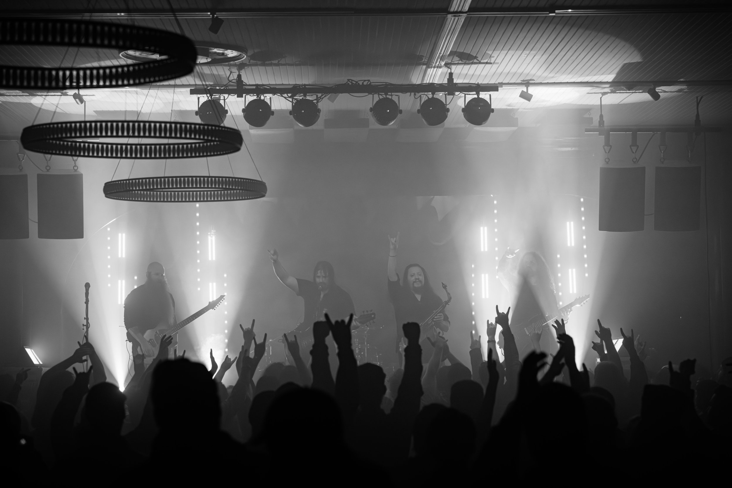 A black and white photo of a live concert with a band performing on stage and an audience raising their hands, many making the rock on gesture.