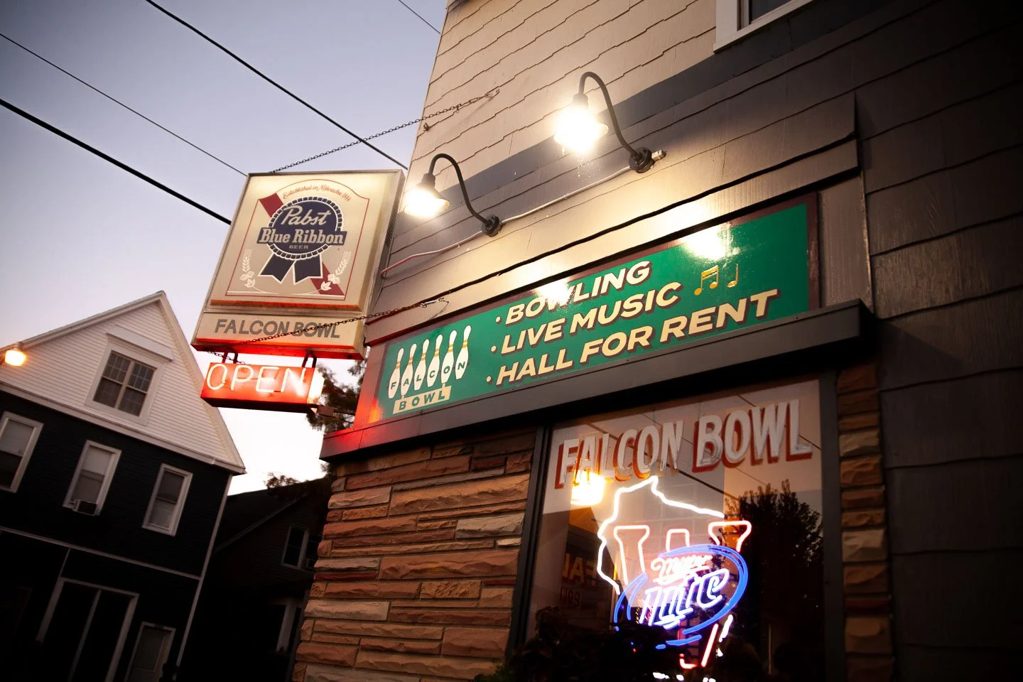 The exterior of Falcon Bowl features a sign advertising bowling, live music, and a hall for rent. Neon signs include a Pabst Blue Ribbon beer sign and a neon 'Miller Lite' sign. The building has brick and wooden siding. There is a nearby house with white siding and multiple windows. The sky is at dusk.