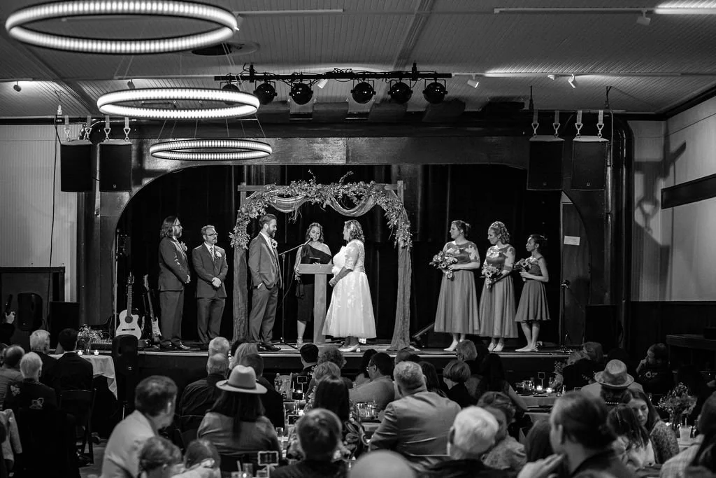 A black and white photo of a wedding ceremony on stage with seven people present, surrounded by an audience in a banquet hall with chandeliers and stage lighting.
