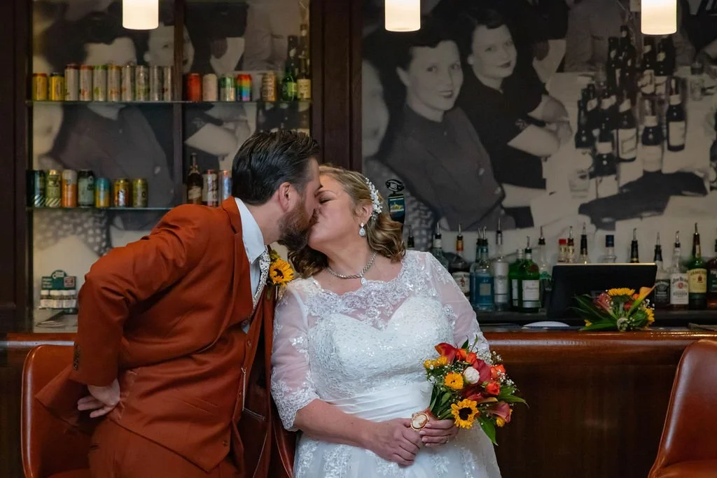 A bride and groom share a kiss at a wedding reception in a bar, with a black-and-white mural of women and bottles of alcohol behind them.
