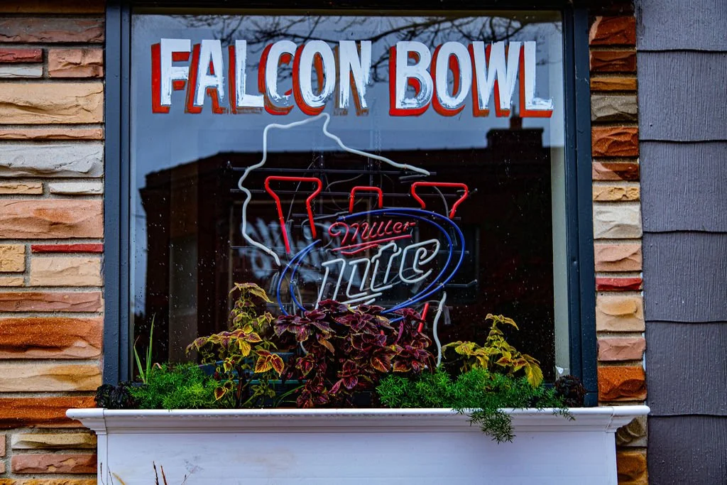 Sign in front of a window with a neon sign that reads "Miller Lite" and the words "Falcon Bowl" above it, surrounded by a planter with various plants.