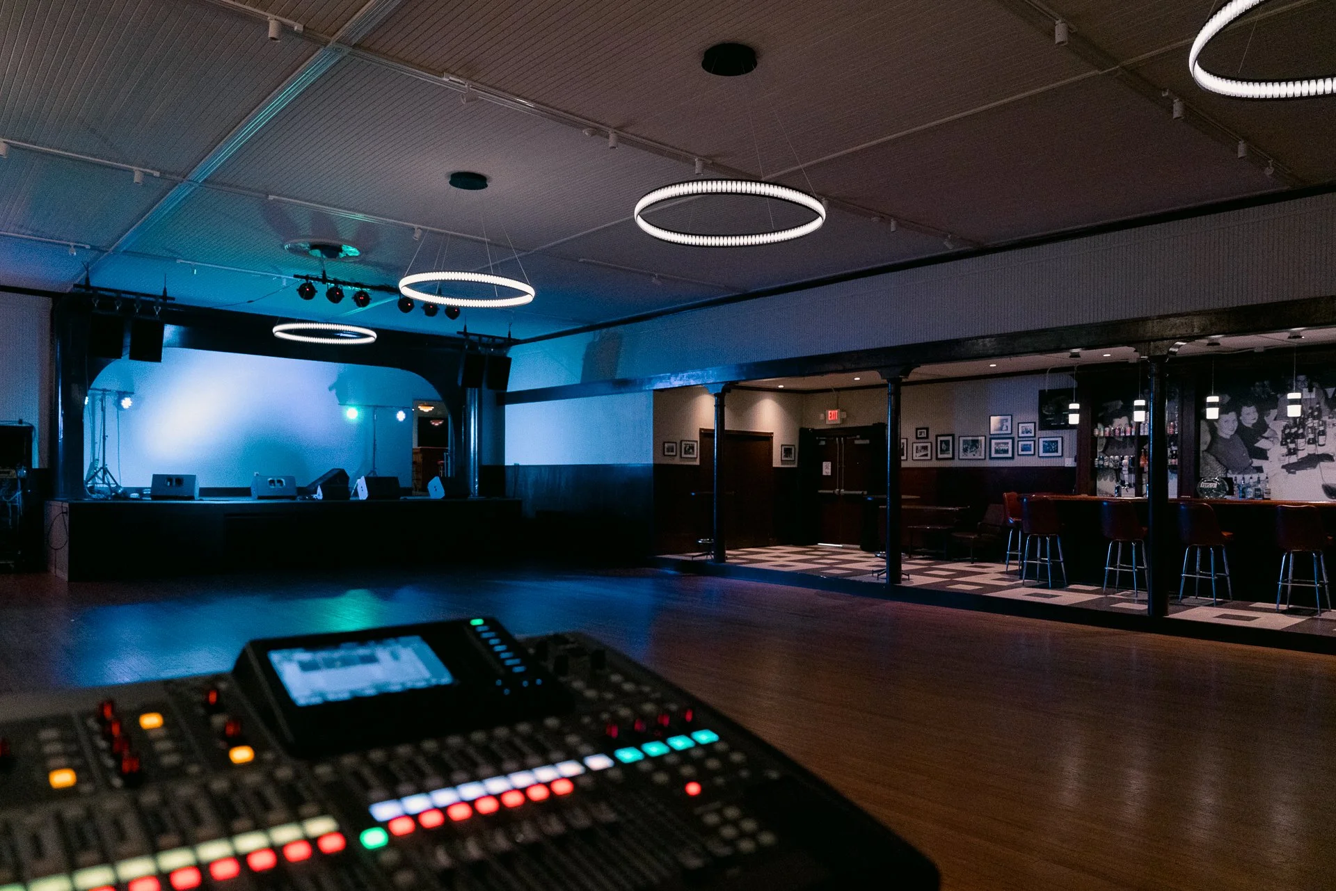 An empty stage with sound and lighting equipment and a dimly lit party area with bar stools and artwork on the wall.
