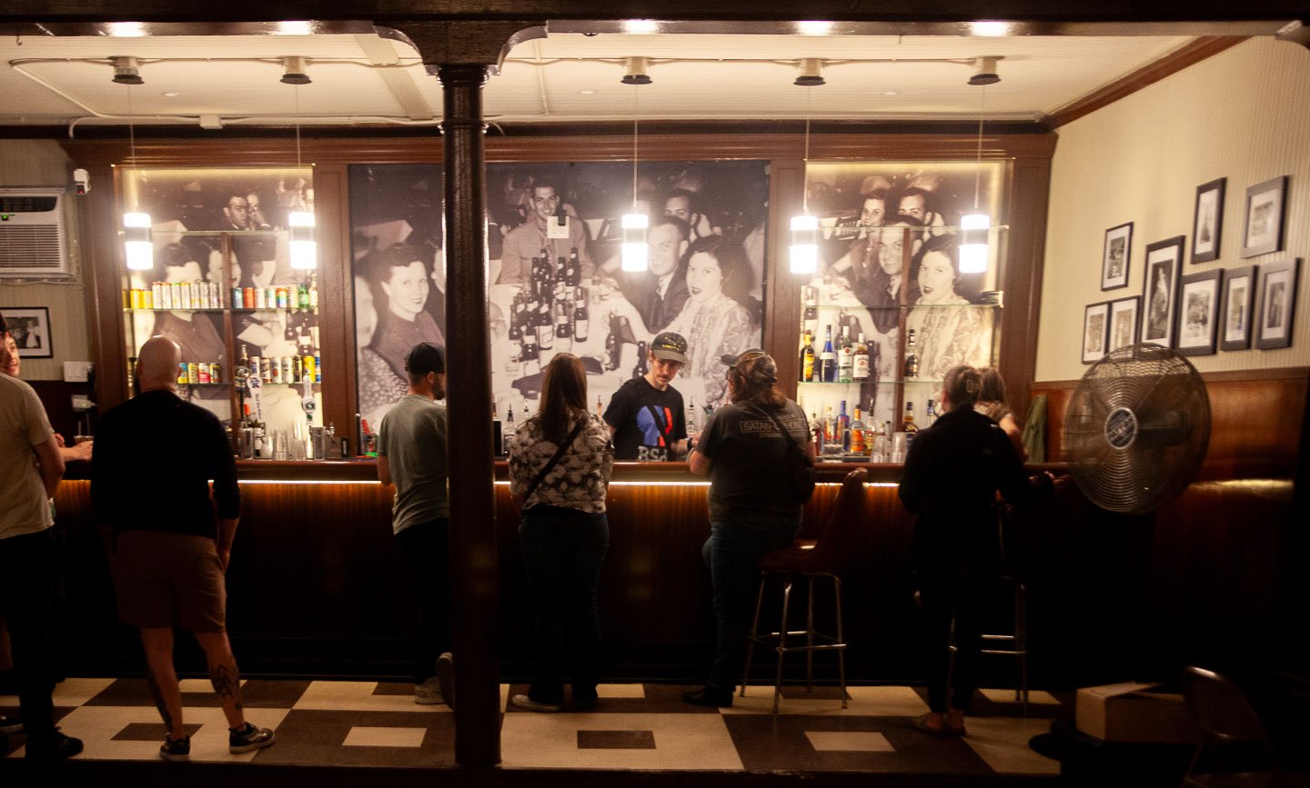 Bar with patrons standing and sitting, bartender behind the counter, vintage black-and-white photographs on the wall, framed pictures on the right wall, checkered floor, large fan to the right.