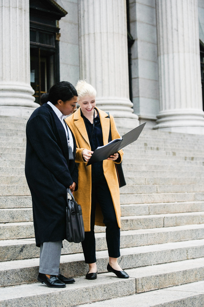 Two women standing on steps in front of a government building, looking at a binder or folder and discussing, one in a dark coat and the other in a tan coat.