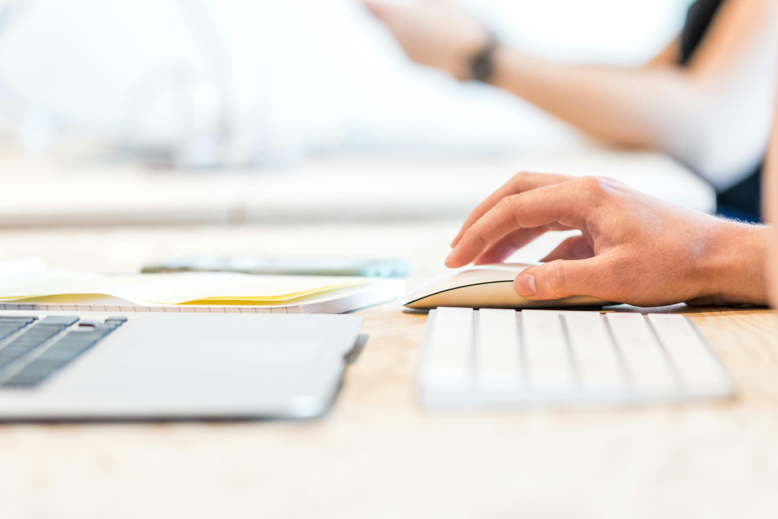 Close-up of a person's hand using a computer mouse at a desk with a laptop, keyboard, and notepad.
