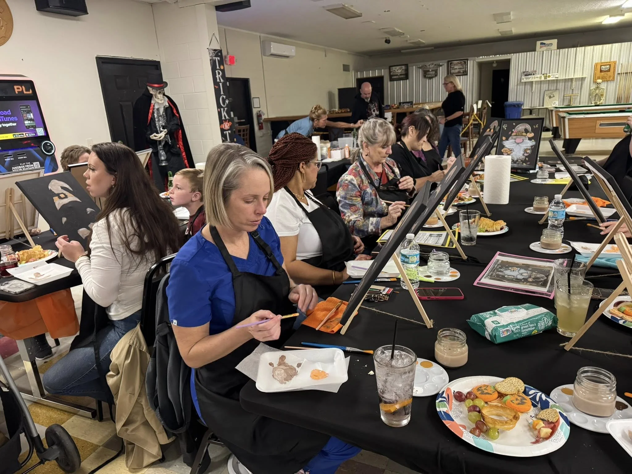 People participating in a group art event painting portraits on canvases at a long table decorated with Halloween-themed cookies and snacks in a community room.