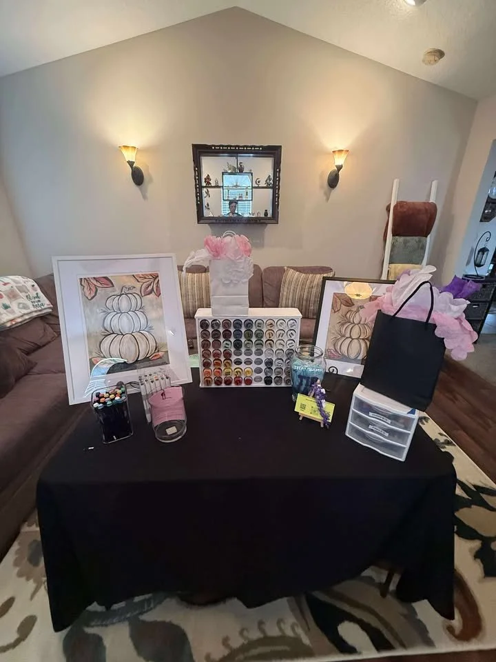 Table with art supplies, framed pumpkin artwork, a black gift bag, and decorative items, in a living room with couches and wall sconces.