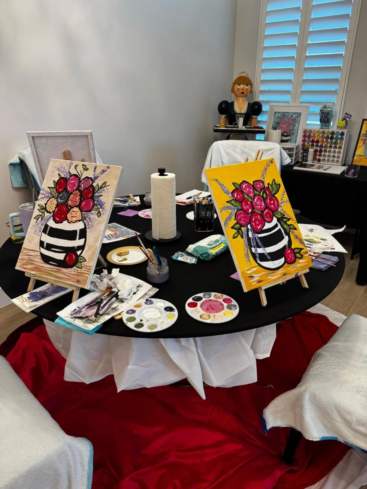 An art studio with two small, colorful still life paintings of vases with flowers on easels, surrounded by painting supplies on a black round table, covering a white tablecloth, with a red fabric draped underneath. In the background, there are shelves with art supplies, a mannequin head, and a window with white shutters.