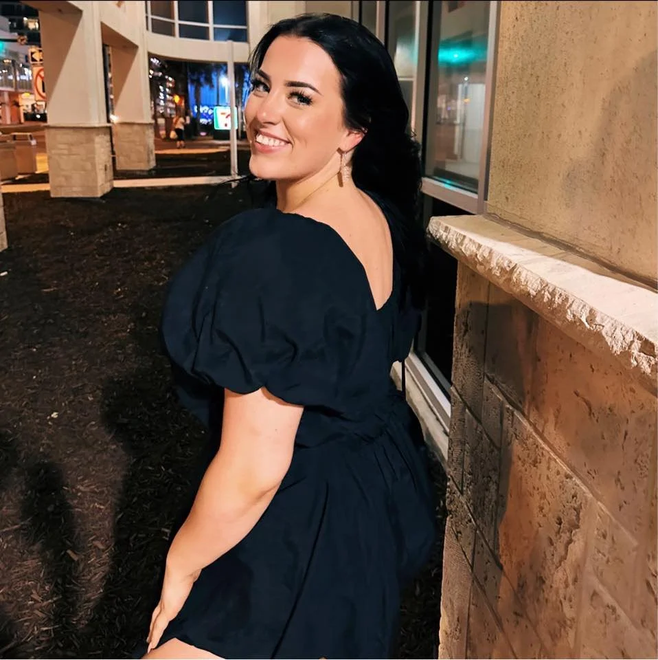 A woman with dark hair smiling at night outside a building, wearing a navy blue dress with puffed sleeves and a V-neck, standing near a stone wall.