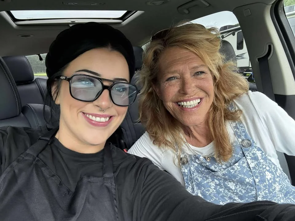 Two women sitting inside a car, smiling at the camera for a selfie. One has black hair, glasses, and a septum piercing, wearing a black top, and the other has red hair, freckles, wearing a white shirt and a blue floral apron.