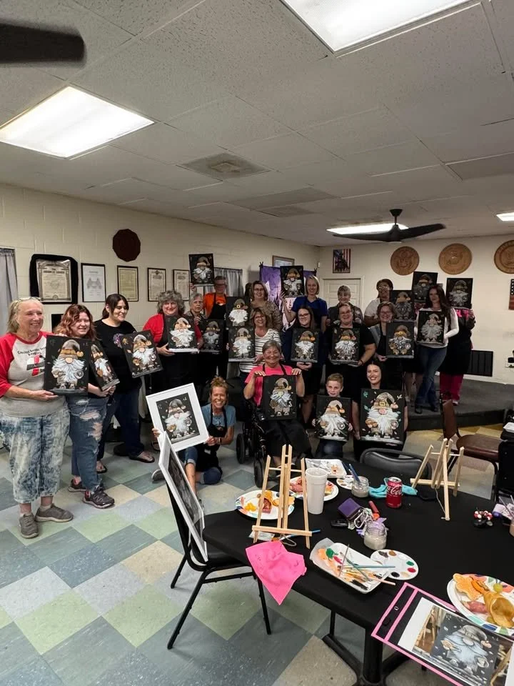 Group of women in an art class holding up paintings of Santa Claus as they smile for the photo in an art studio or classroom.