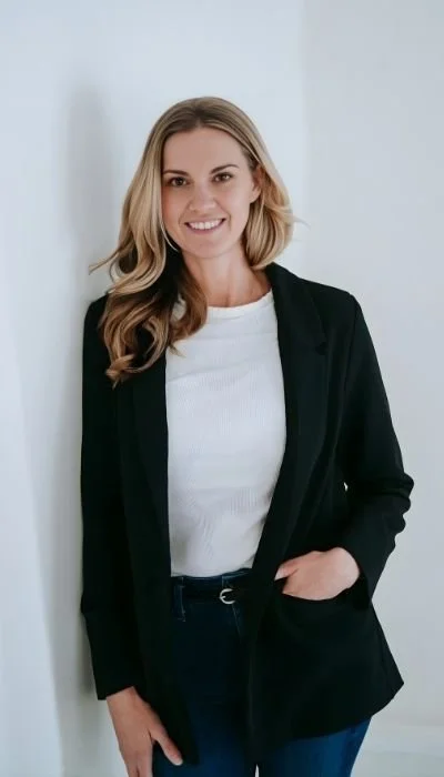 A woman with shoulder-length blonde hair, wearing a white T-shirt and black blazer, standing against a white wall and smiling.