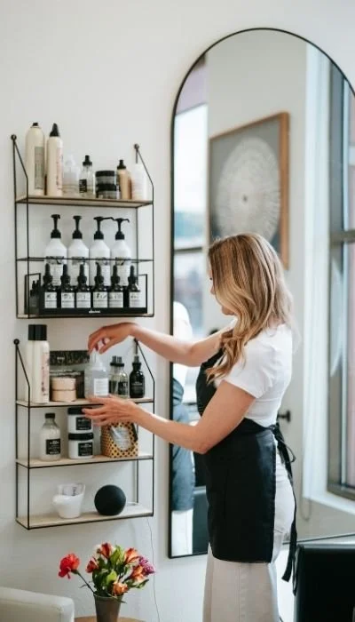 Woman organizing haircare products on a black metal shelf in a bright hair salon with a large mirror.