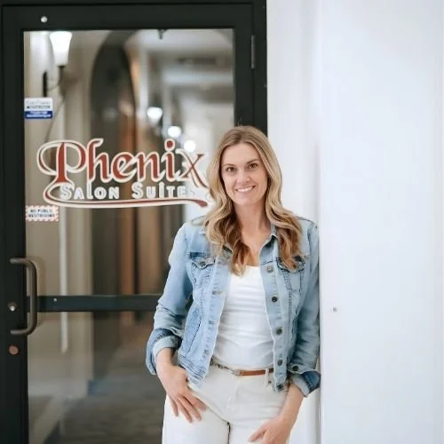 A smiling woman stands in front of a salon door labeled 'Phenix Salon Suites' in a building corridor in Colorado Springs.