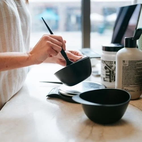 A hairstylist stirring a black bowl with hair color in a hair salon. Salon items, including another empty black bowl, a large bottle, and a container in a cup with a patterned sleeve, are on the counter.