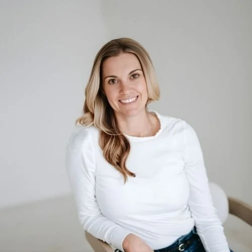 Chelsea Villari, a hairstylist with long blonde hair, smiling while sitting on a beige chair against a plain light gray background.