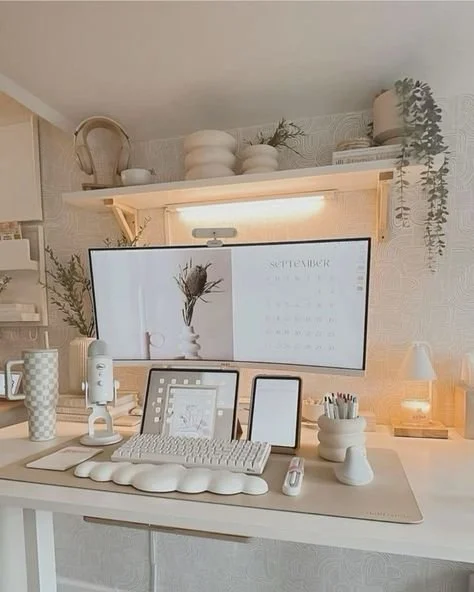 A tidy home office desk with a large computer monitor displaying a September calendar and a flower illustration, a tablet, a smartphone, a keyboard, a microphone, and various stationery items, with wall shelves holding plants, vases, and decorative items in a cozy, well-lit room.