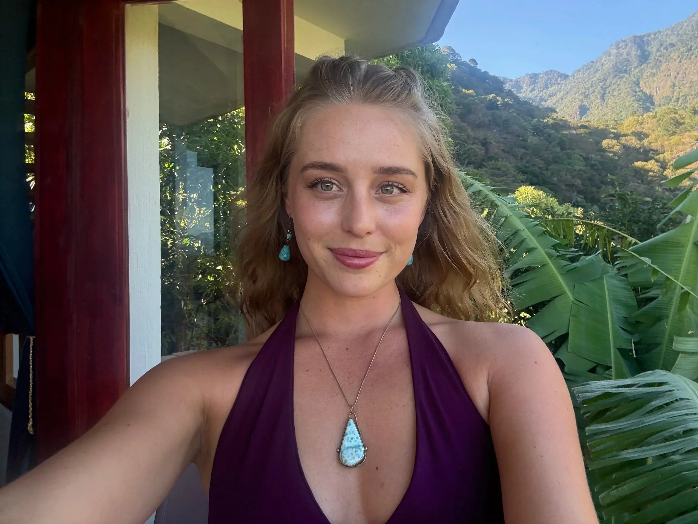 A young woman with curly blonde hair, wearing a purple halter top and turquoise jewelry, is taking a selfie outdoors with green mountains and lush vegetation in the background.