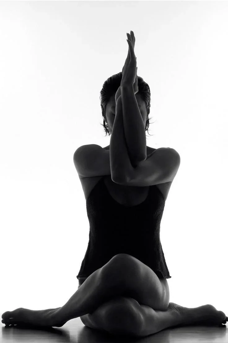 A person practicing yoga in a seated pose with legs crossed, one arm bent and raised over her head, and the other arm crossing in front of her face, in black and white.