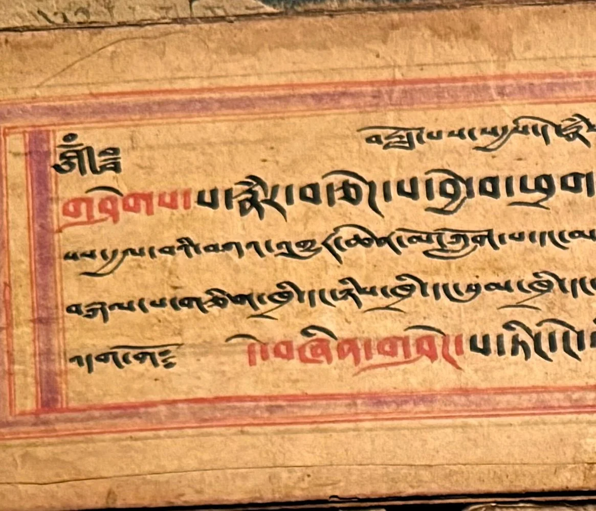 Close-up photo of an ancient manuscript with text in a script resembling Sanskrit or Tibetan, written in black and red ink, on aged paper with a decorative border.