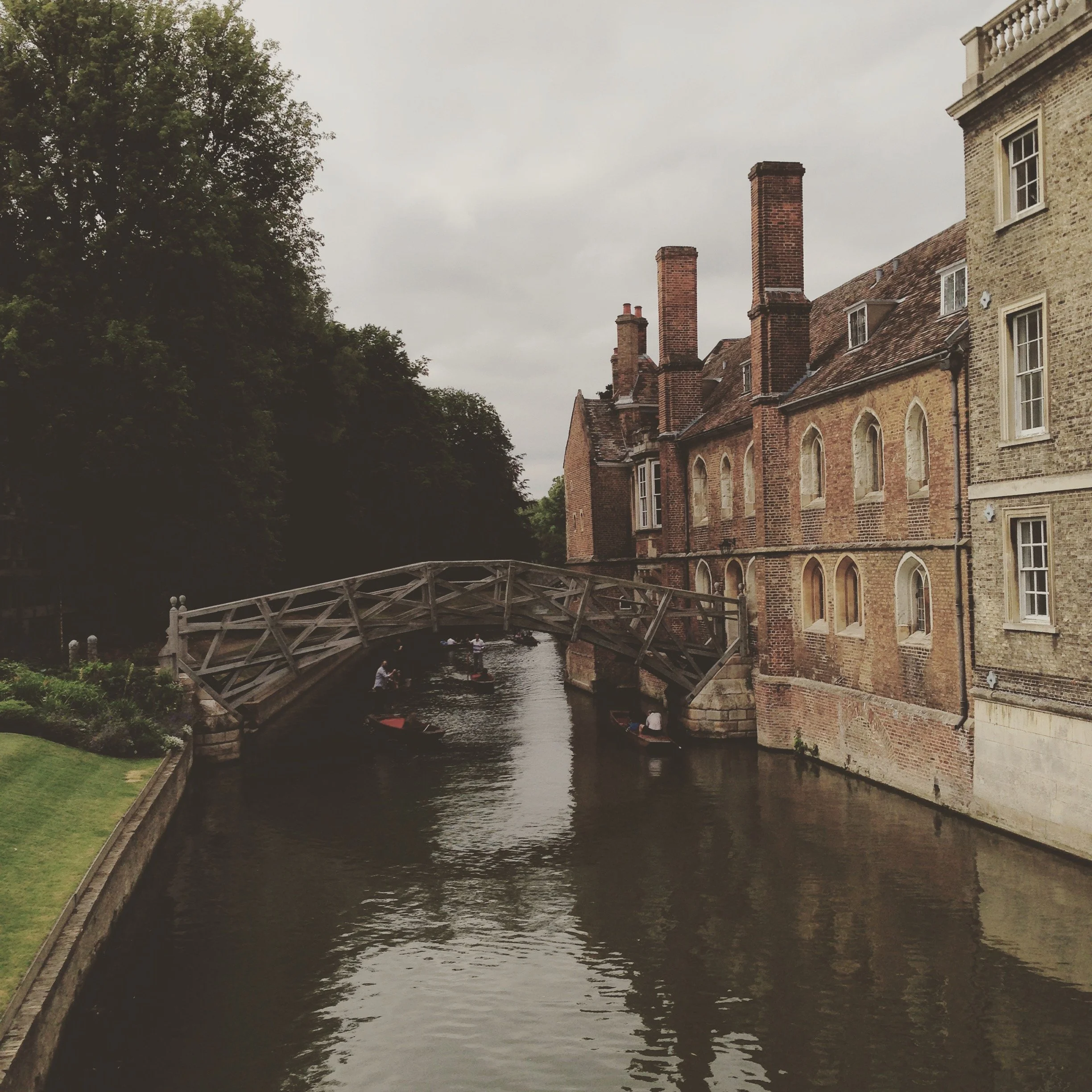 A historic brick building by a canal with a small wooden bridge and people riding boats, surrounded by trees and overcast sky.