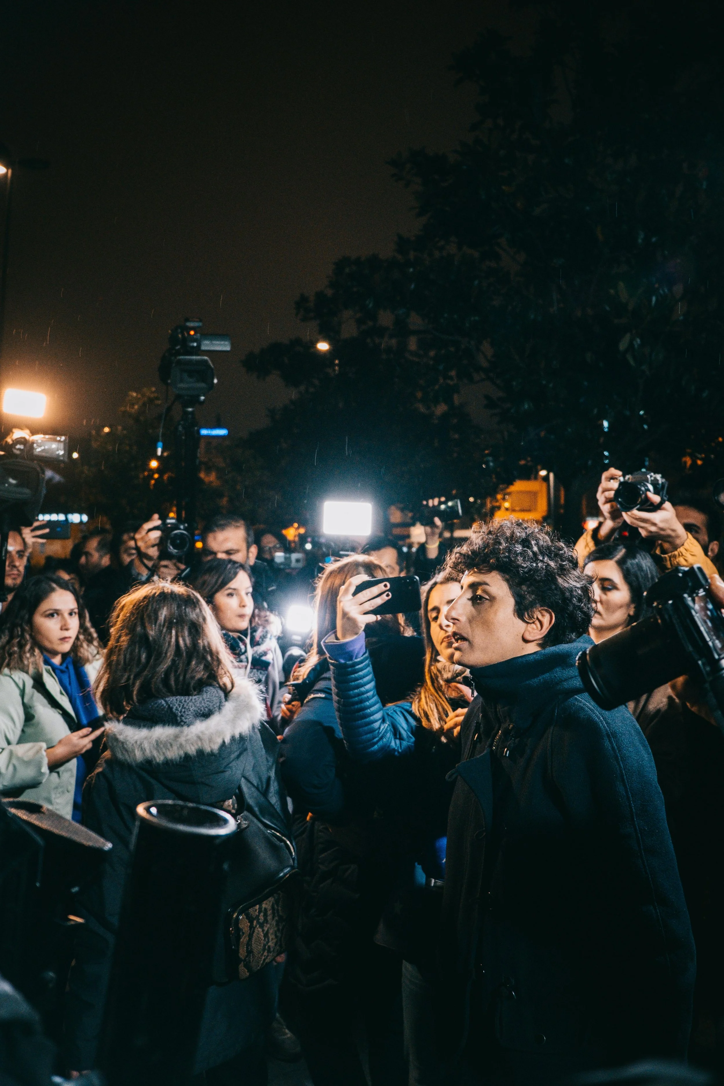 A crowd of people, some holding cameras and phones, gathered outdoors at night, with bright lights and trees in the background.